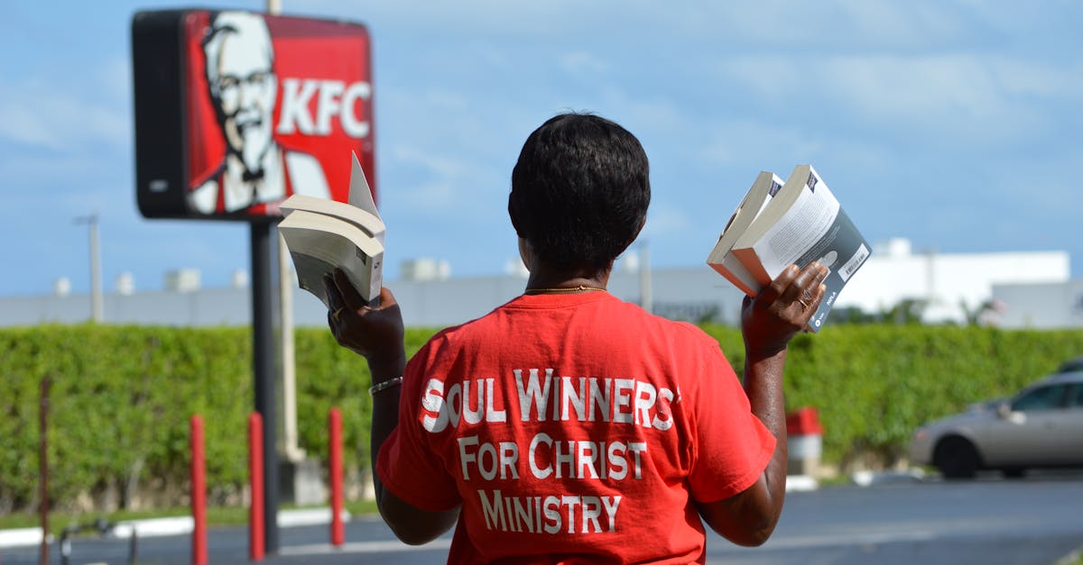A street preacher in a red t-shirt holds books outside a KFC, advancing their message.